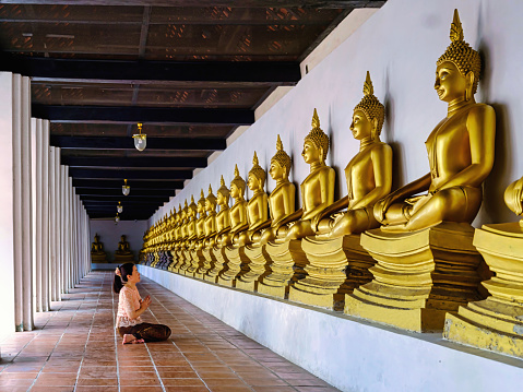 Asian woman in traditional dress sitting on floor pray and worship to Golden Buddha statue that lined up at Wat Phutthaisawan, Ayutthaya, Thailand