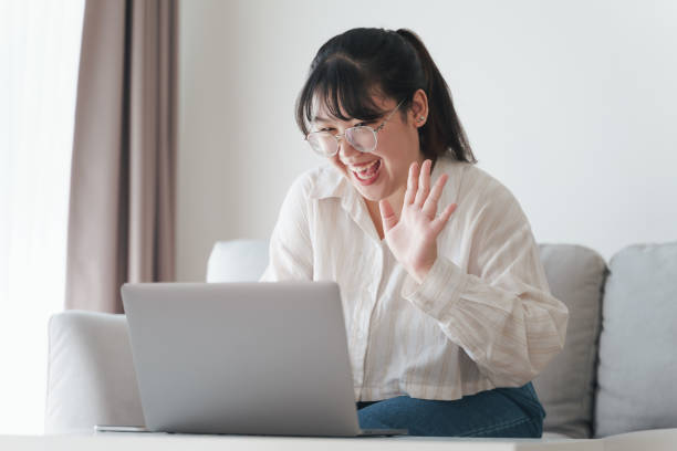 istock Young Asian woman using laptop computer for online video conference call waving hand making hello gesture on the couch in living room. 1397670108