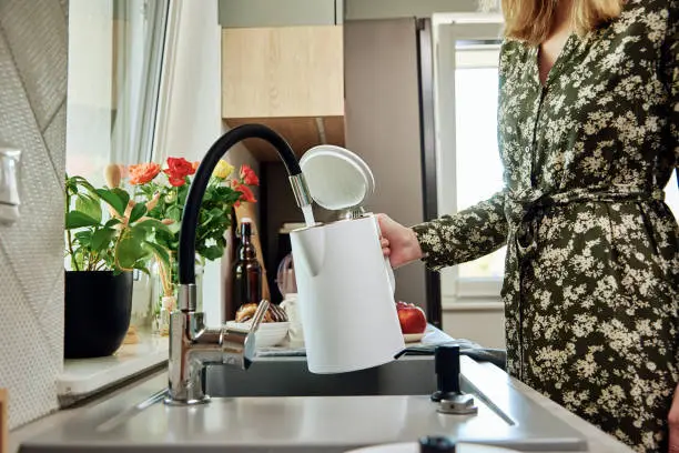Woman pouring clean filtered water from faucet into electric kettle for boiling water at kitchen