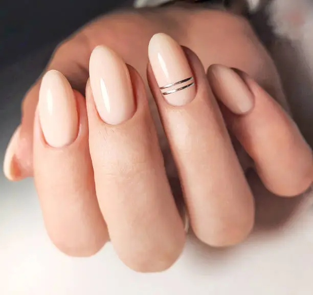 Gentle gel polish of beige color on almond-shaped nails with silver stripes. Nude manicure with shiny stripes design. Pastel color coating on the nails.
