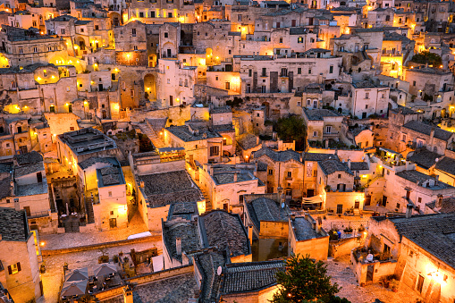 An idyllic cityscape at twilight of the maze of alleys and stone houses in the old town of Matera, known worldwide as the 'Sassi di Matera' (Matera's Stones Hills). In the image the Sasso Barisano hill, seen from a belvedere. The ancient city of Matera, in the region of Basilicata, in southern Italy, is one of the oldest urban settlements in the world, with a human presence that dates back to more than 9,000 years ago, in the Paleolithic period. The Matera settlement stands on two rocky limestone hills called 'Sassi' (Sasso Caveoso and Sasso Barisano), where the first human communities lived in the caves of the area. The rock cavities have served over the centuries as a primitive dwelling, foundations and material for the construction of houses, roads and beautiful churches, making Matera a unique city in the world. In 1993 the Sassi of Matera were declared a World Heritage Site by Unesco. Super wide angle image in high definition format.