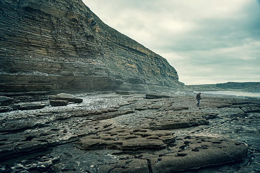 A lone female beachcomber walks a rocky beach overlooked by high cliffs and a stormy sky.