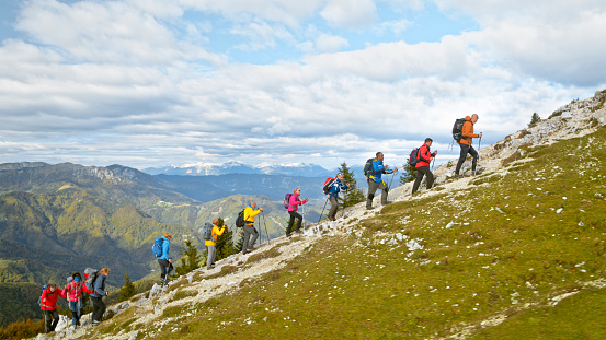 Group of hikers with hiking poles and rucksacks hiking climbing mountain against cloudy sky.