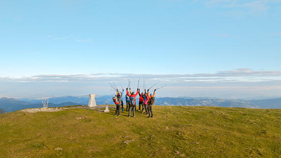 Group of hikers with hiking poles standing on grassy mountain top against sky.