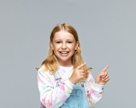 Portrait of smiling cute girl in casuals pointing while standing against gray background