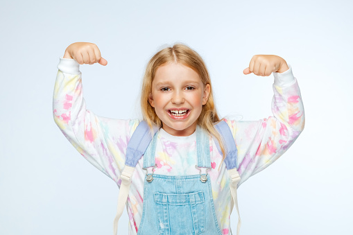Portrait of happy schoolgirl with backpack flexing muscles while standing against white background