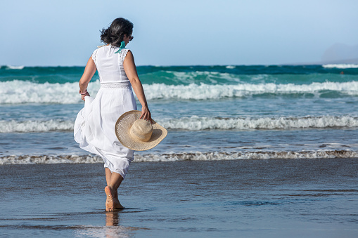 Back view of brunette in white dress with straw hat walking on wet beach towards foamy sea waves on summer day on resort