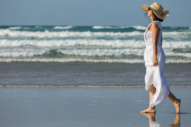 Side view of barefoot female in white maxi dress and straw hat walking on wet sand against sea waves and blue sky while spending weekend day on beach