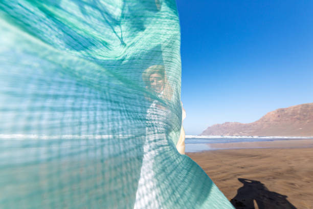Side view of happy woman in white dress looking at camera with smile through transparent scarf while dancing on sandy seashore against cloudless blue sky on weekend day