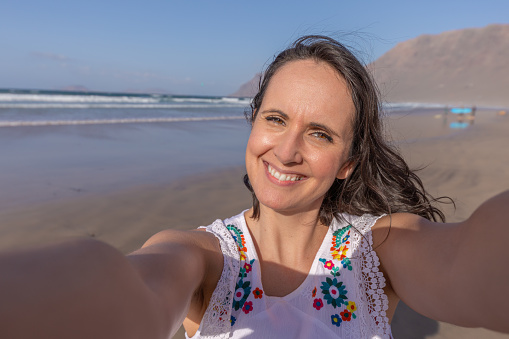 Cheerful middle aged woman with dark hair smiling and looking at camera while taking selfie on sandy beach near waving sea on summer weekend day