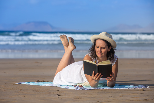 Barefoot middle aged female tourist in white dress and hat relaxing on sandy beach and reading interesting book against waving sea and blue sky on summer weekend day on resort