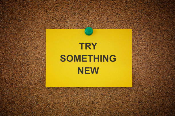 A sticky note with the words Try Something New on it pinned to a cork board. Close up.