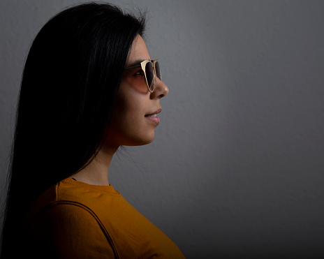 Young lady profile portrait in a studio shot. She is latin in her 30s, has long black hair and is wearing sunglasses