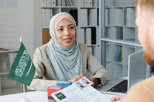 Modern Muslim woman wearing hijab working as consular officer in Saudi Arabia embassy interviewing Caucasian man for visitor visa