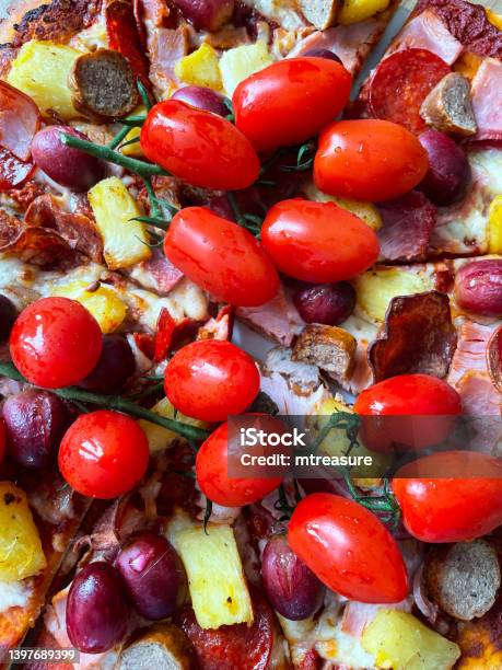 Full Frame Image Of Freshly Baked Sliced Homemade Pizza Mozzarella Cheese Onion Pepperoni Slices Ham Sausage Pineapple Pieces Red Grapes And Red Pesto Sauce Topping Served With On The Vine Cherry Tomatoes Elevated View Stock Photo - Download Image Now