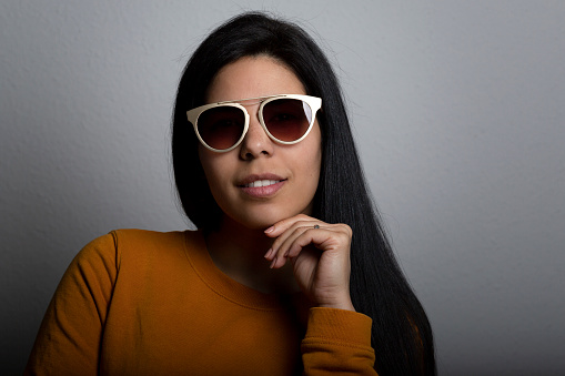 Young lady fashion studio portrait with a hand under her chin holding her face while looking at the camera with sunglasses with a little smile. She is latin in her 30s and has long black hair