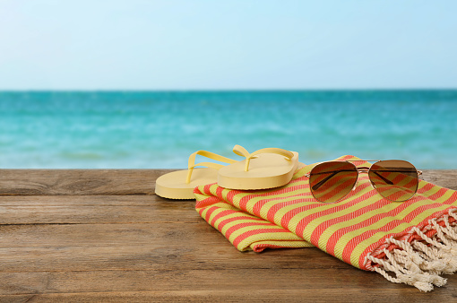 Beach towel, flip flops and sunglasses on wooden surface near seashore. Space for text