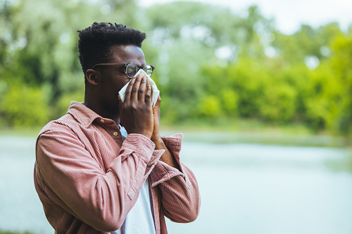 Sick Man With High Temperature is Using Handkerchiefs, Trying to Override the Sickness While Walking in Nature. Allergic black man blowing on wipe in a park on spring season
