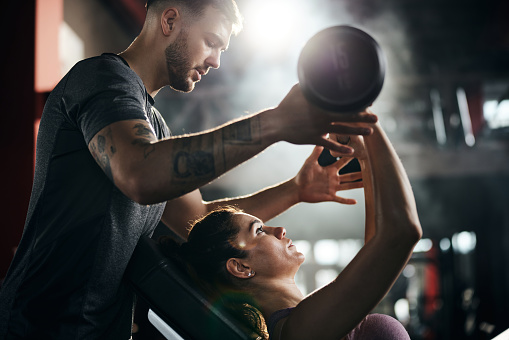 Young man helping his girlfriend during her sports training with barbell in a health club.