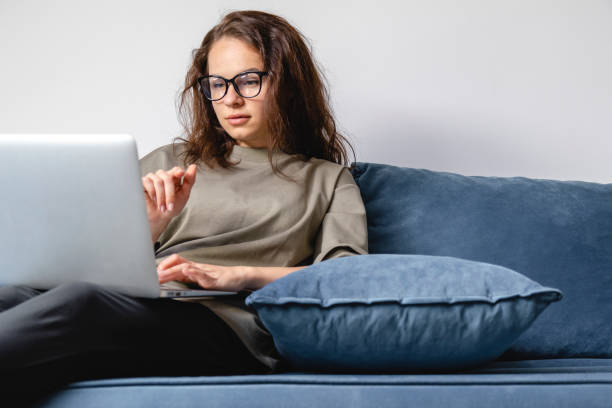 jeune femme aux cheveux bruns en lunettes utilisant un ordinateur portable tout en étant assise sur le canapé à la maison. - women computer home interior brown hair photos et images de collection