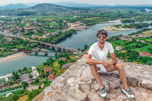 Young handsome European man sits on the wall of Rozafa Castle on the backdrop of Drin river valley with bridges and Bahcallek village in Albania. Guy poses on the backdrop of a beautiful landscape