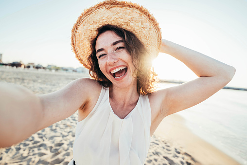 Young joyful woman in white shirt wearing hat smiling at camera on the beach - Traveler girl enjoying freedom taking selfie on a sunny day - Wellbeing, healthy lifestyle and happy people concept