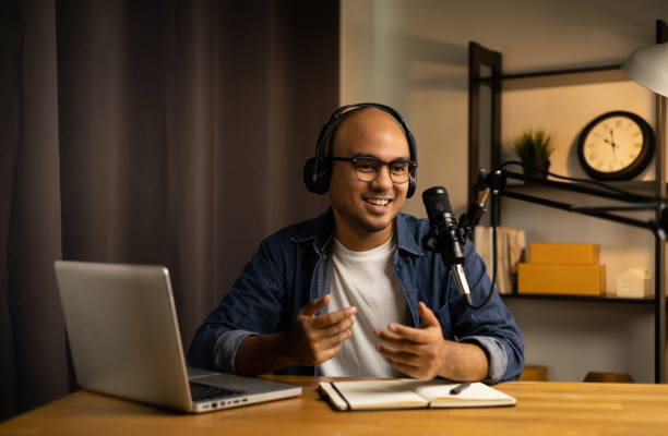 istock Young asian man host streaming podcast with condenser microphone work on laptop at small broadcast home studio. Content creator blogger recording voice over radio interview guest conversation 1397702110