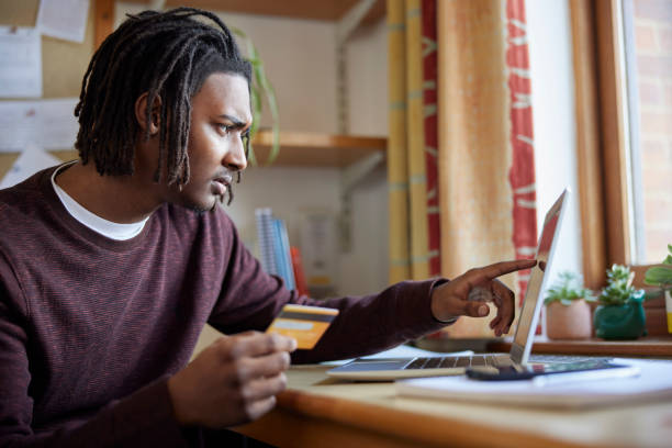 männlicher universitäts- oder college-student mit kreditkarte, der auf einen laptop schaut, besorgt über schulden am schreibtisch im zimmer - student loans stock-fotos und bilder