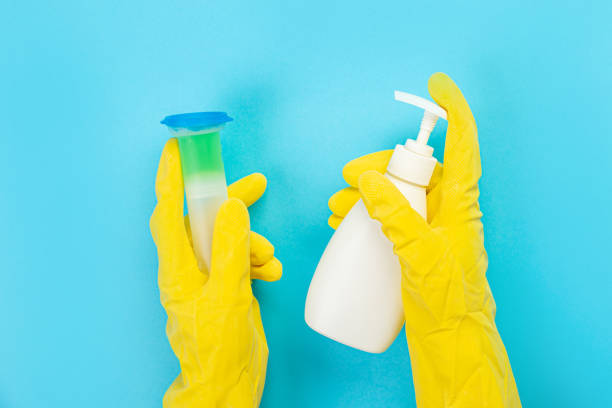 The hands of a housewife in yellow rubber protective gloves hold a bottle of household chemicals and a rag. Detergent for various surfaces in the kitchen, bathroom and other rooms. Light pastel blue background. las manos de un ama de casa con guantes protectores de goma amarilla sostienen una botella de productos químicos domésticos y un trapo. detergente para diversas superficies en la cocina, baño y otras habitaciones. fondo azul pastel claro.