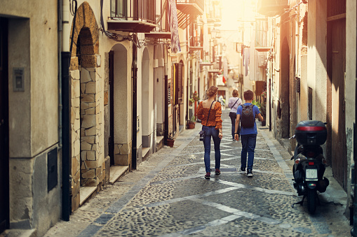 Tourists walking in beautiful narrow streets of Italian town of Cefalu. Sunny summer day in Italy, Sicily.\nWoman and two teenagers are walking in the street.\nCanon R5