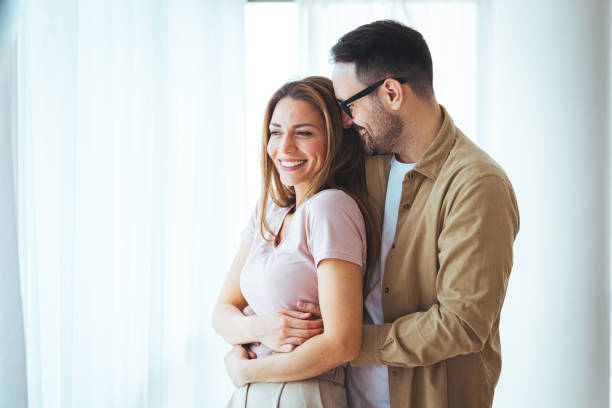 istock Smiling couple hugging each other and standing near window while looking outside. 1397716297
