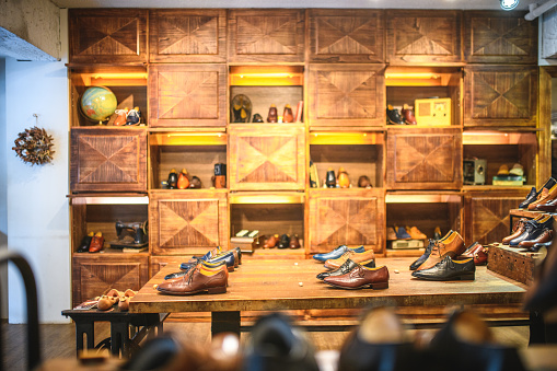Elegant designer shoe store for gentlemen with wooden shelves and warm lightening. Display of stylish leather shoes, boots, Oxford shoes, brogue shoes and casual footwear.