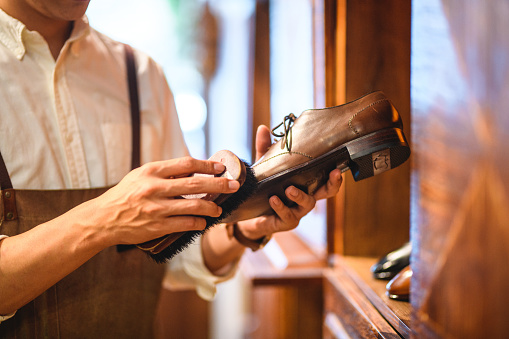 Caucasian salesman cleaning and polishing fancy leather shoes in a footwear store. He is holding a brown shoe with one hand and with the other he is brushing it and making it shiny. He is wearing a white shirt and an apron.