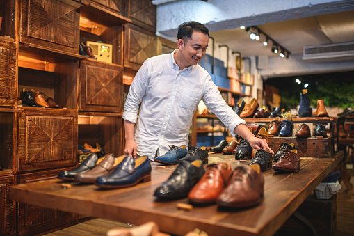 Asian businessman choosing discounted high quality elegant leather shoes. He likes to wear stylish shoes and look elegant at work. Leather shoes on wooden shelves in the background and foreground.
