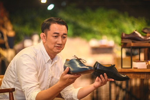 Chinese male adult deciding which leather shoes to buy. He is holding a blue and a black shoe on each hand. He is stressed because he doesn't know which one to take home.