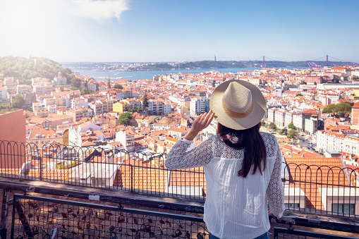 A tourist woman on a sightseeing trip overlooks the colorful cityscape of Lisbon, Portugal, with Alfama district, castle Sao Jorge and 25th of April bridge