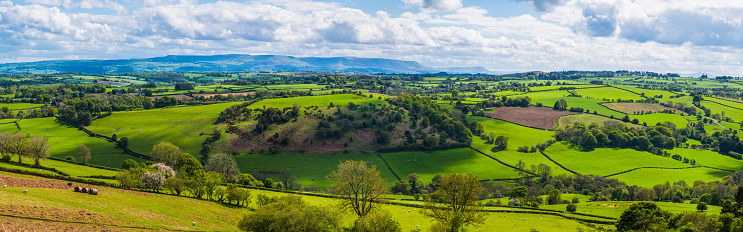 Panoramic view across idyllic patchwork landscape of green fields, farmland and pasture to mountains beyond.