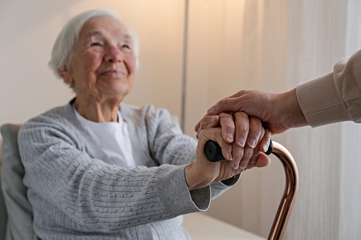 Senior woman holding quad cane handle in elderly care facility. Unrecognizable female comforting mature lady in nursing home, sitting with walking stick. Background, close up, cropped shot.