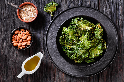 kale chips in black bowl with almonds, sesame seeds, olive oil on dark oak textured table, horizontal view from above, flat lay