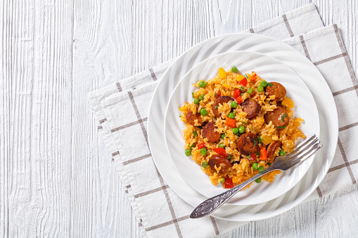 browned smoked sausages with rice, bell pepper, onion and green peason white plate on white wooden table with napkin and fork, horizontal view from above, flat lay, free space