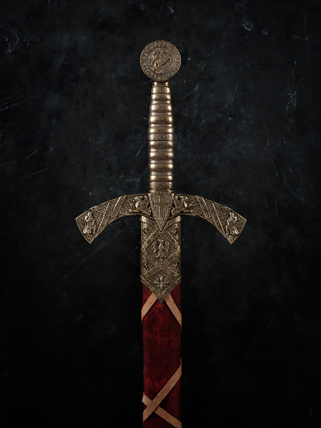 Vertical closeup of a knight's sword with a bronze handle, against a dark background.