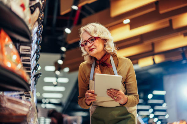 Senior female employee using tablet in market Senior woman working at the supermarket wearing apron and glasses and checking shelf with groceries working seniors stock pictures, royalty-free photos & images