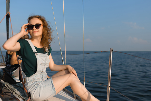 A young woman wearing stylish sunglasses is sitting on a yacht. The woman on summer vacation enjoying the view of the sea