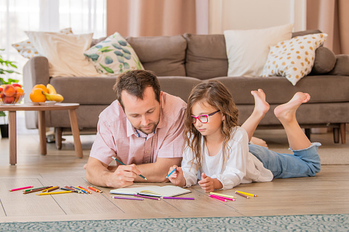 Father and daughter in glasses are drawing together while lying on the floor in the apartment room.