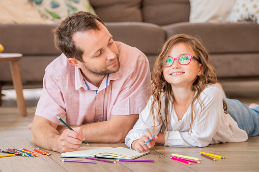Father and daughter in glasses are drawing together while lying on the floor in the apartment room.