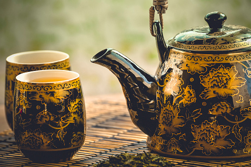 Golden and black teapot and two tea cups on the straw table, with green tea and soft green nature background, on Japanese tea ritual.