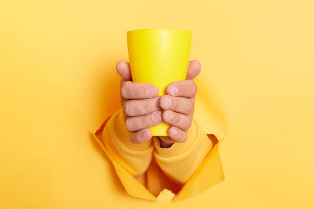Cropped view of man holding coffee to in mug in both hands through hole in paper wall isolated on yellow background.