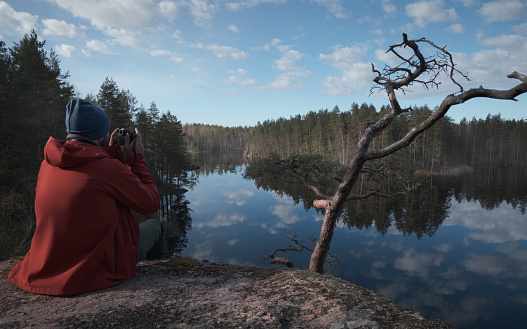 A young person on the shore of a mirror picturesque forest lake. A man photographs a picturesque landscape. Back view