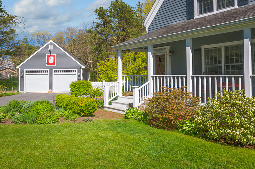 The covered front porch and detached garage of a small single family home  on Cape Cod on a springtime afternoon with a green lawn and shrubbery