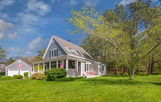 A small single family home with a detached two car garage on Cape Cod on a springtime afternoon with a green lawn and backyard trees.